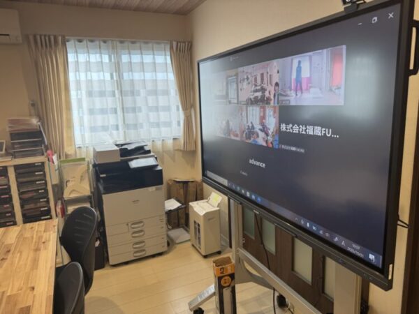 部屋の中に大型モニターが設置され、オンライン会議の画面が映っている。 画面には複数の施設や人の映像が分割表示され、スタッフが活動している様子が見える。 左側にはコピー機や書類棚、木目のデスクと黒い椅子があり、オフィスのような環境。 窓からは自然光が入り、白いカーテンがやわらかく光を通している。 福祉施設の拠点同士がオンラインでつながり、交流や会議をしている様子を写した写真。