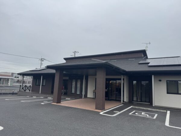 曇り空の下、平屋建ての建物が正面から写っている。 建物は黒い屋根と白い外壁で、玄関前には茶色の柱と屋根付きの入り口がある。 屋根の一部には太陽光パネルが設置されており、駐車場には車いすマークの専用スペースが描かれている。 全体的に落ち着いた雰囲気で、福祉施設やデイサービスなどを思わせる外観の写真。