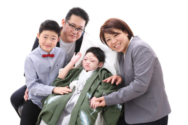 白い背景の前で、4人の家族が並んで写真に写っている。 中央には緑の羽織と白い着物を着た子どもが座っており、両脇から家族がやさしく寄り添っている。 左側には赤い蝶ネクタイをつけた男の子、右側にはグレーのスーツを着た女性が笑顔で手を握っている。 後ろには黒縁の眼鏡をかけた男性が優しく見守るように立っている。 家族全員が温かな笑顔で、絆と愛情が伝わる記念写真。