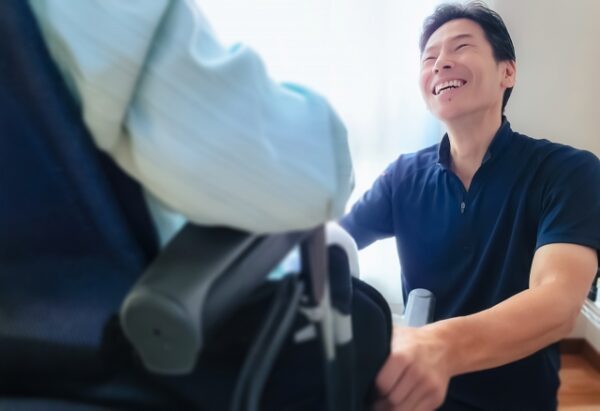 車いすに座る人と、介助をする男性が室内で向かい合っている。 男性は濃い青色のポロシャツを着ており、やさしい笑顔で相手に話しかけている。 男性は片手で車いすのひじ掛けあたりを支えながら、もう片方の手を膝の上に置いている。 背景には白いカーテンから柔らかい光が差し込み、全体的に温かく穏やかな雰囲気の写真。