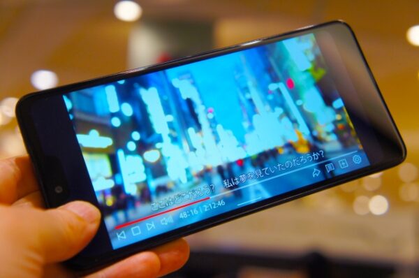 手にしたスマートフォンの画面に、字幕付きの映像が映し出されている。 街の灯りがぼんやりと輝き、バリアフリー字幕を通じて物語の世界が広がっていく。