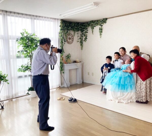 フォトスタジオで、カメラマンが家族の写真を撮影している。 青いドレスを着た子どもを中心に、家族が寄り添いながら笑顔を見せる、あたたかな一場面。