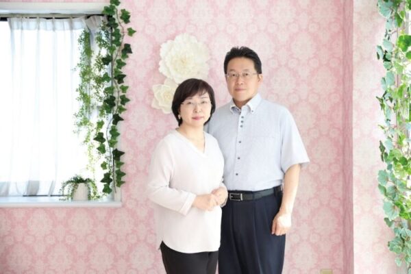 ピンクの花柄の壁の前で、夫婦が穏やかに並び立ち、やさしく微笑んでいる。 やわらかな光に包まれた空間が、安心とあたたかさを感じさせる一枚。