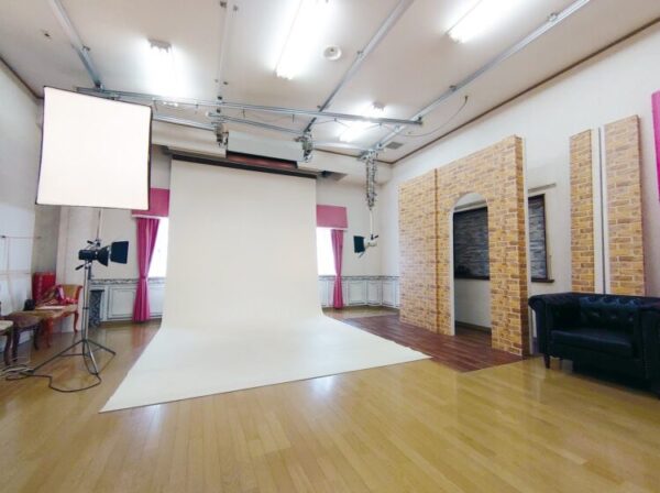 明るい照明に照らされたフォトスタジオの内部。 白い背景紙と撮影用ライト、レンガ調のセットが整えられ、温かみのある空間が広がっている。