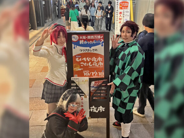 夜の街中で、コスプレ姿の3人が立て看板の前に並び、笑顔でポーズを取っている。 行き交う人の中で、楽しさと表現する喜びが自然に伝わってくる一場面。