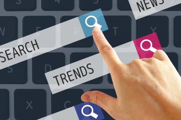 キーボードの上で指が検索アイコンに触れ、「SEARCH」や「TRENDS」などの文字が重なるイメージ。 必要な情報を探し、知ろうとする動きが伝わる一場面。