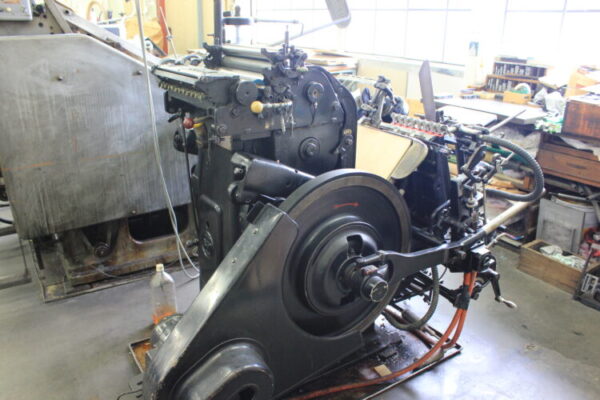 工場内に置かれた、60年以上前のドイツ製の活版印刷機。黒い金属製で、大きなはずみ車の付いた年季の入った機械。